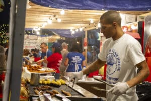 New York City, United States of America - September 22, 2019: Vendor wearing gloves grills corn on the cob and skewers of meat at a food stall during the annual Feast of San Gennaro festival in Little Italy, New York City